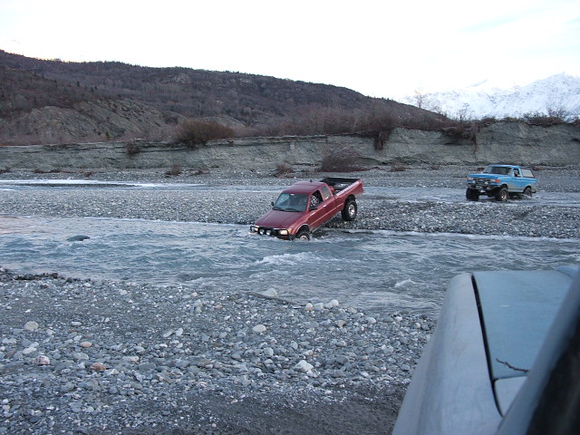 Red compact 4x4 pickup fording a river with a lifted blue truck on the gravel bar
