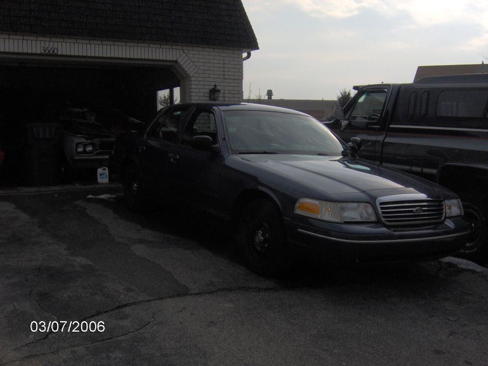 Dark blue Ford Crown Victoria sedan with black steel wheels parked in a driveway next to a garage and pickup truck