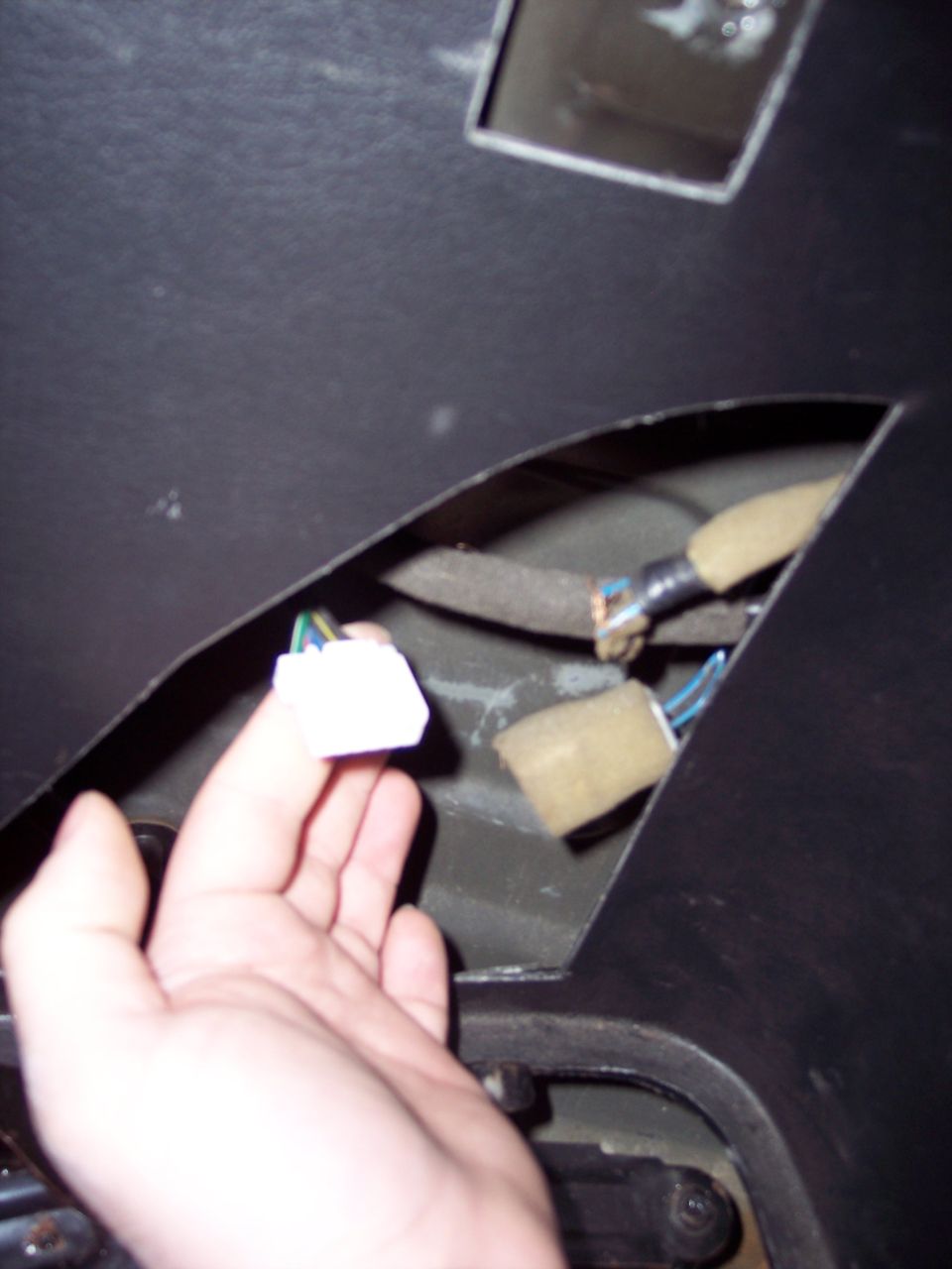 Hand holding a white wiring harness connector near exposed wires and foam padding inside a dark panel