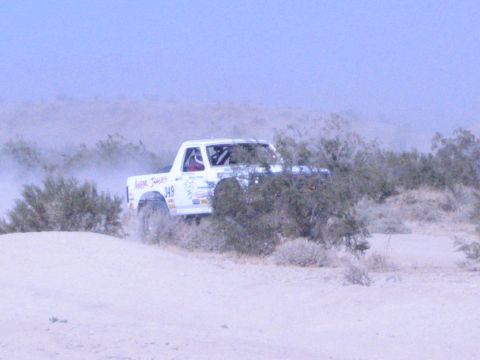 White 949 race truck behind desert shrubs kicking up dust