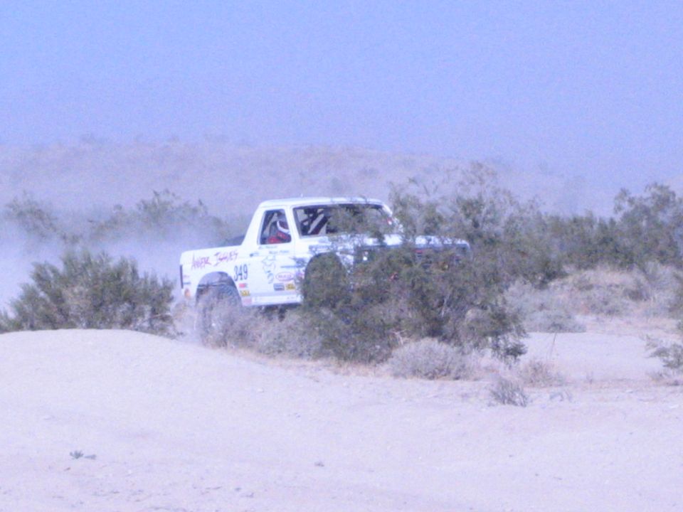 White 949 race truck behind desert shrubs kicking up dust