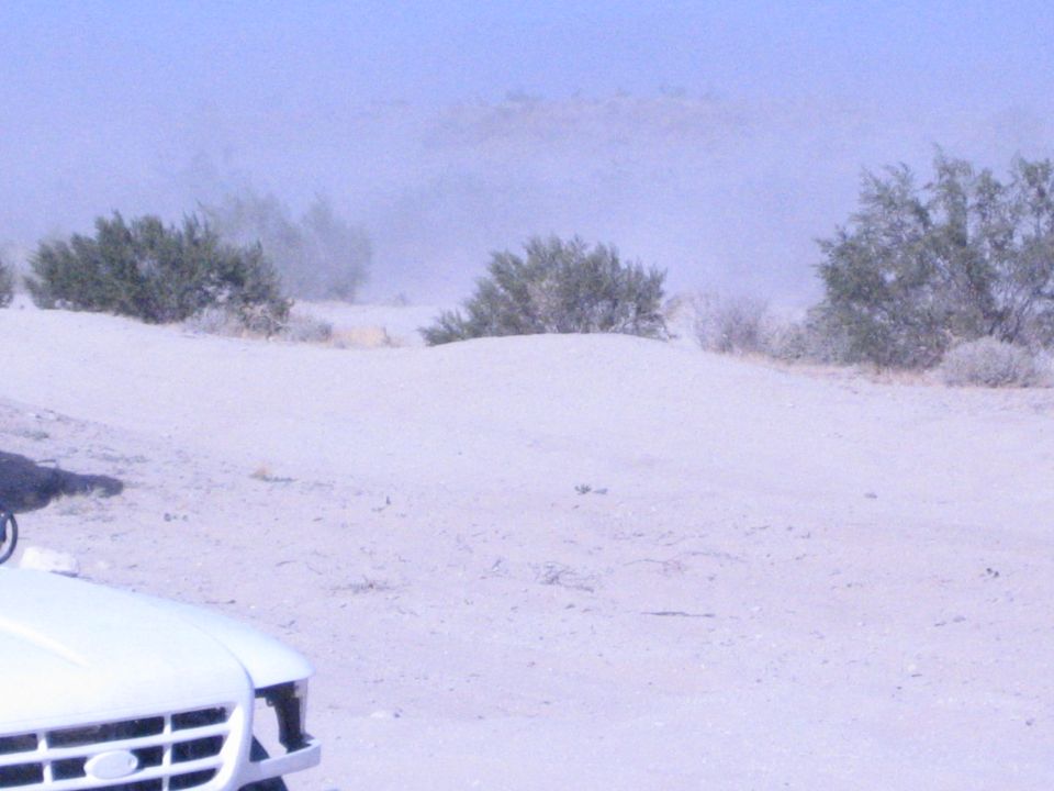 White truck hood at edge of frame on dusty desert flat with brush and haze