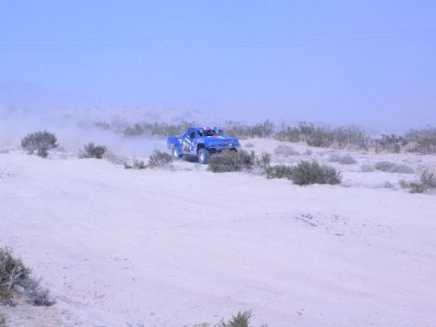 Blue off-road race truck driving along a sandy desert ridge trailing dust