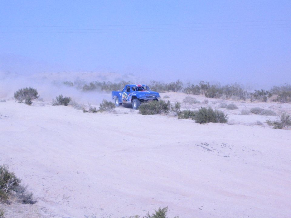 Blue off-road race truck driving along a sandy desert ridge trailing dust