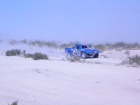 Blue off-road race truck driving through brushy desert flat trailing dust