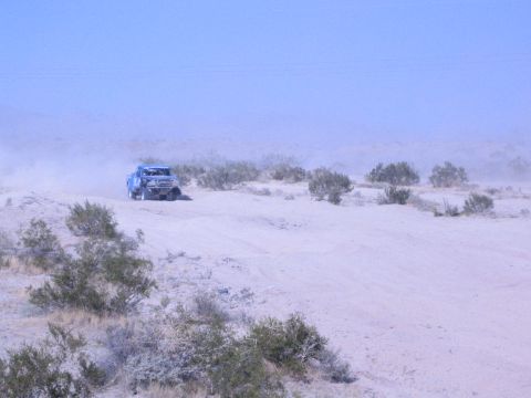Blue off-road race truck in the distance crossing brushy desert in haze
