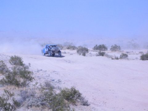 Blue off-road race truck driving across pale desert sand trailing dust