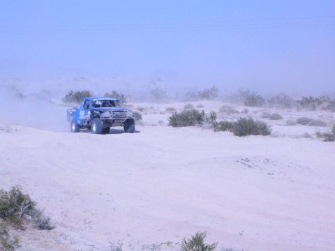 Blue off-road race truck driving across sandy desert flat with dust trail