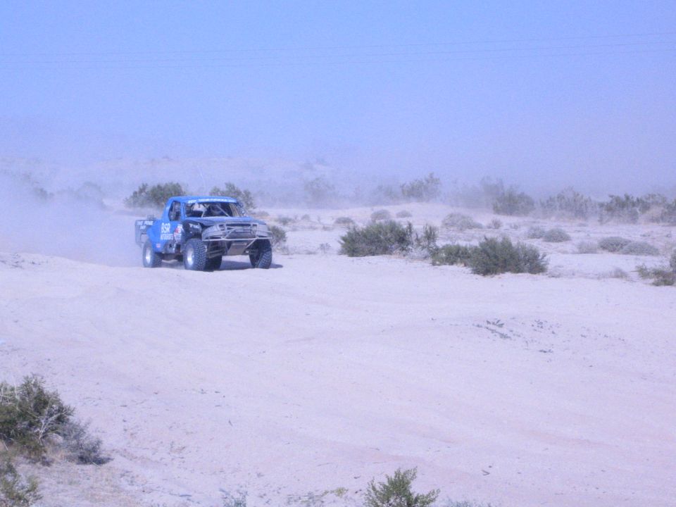 Blue off-road race truck driving across sandy desert flat with dust trail