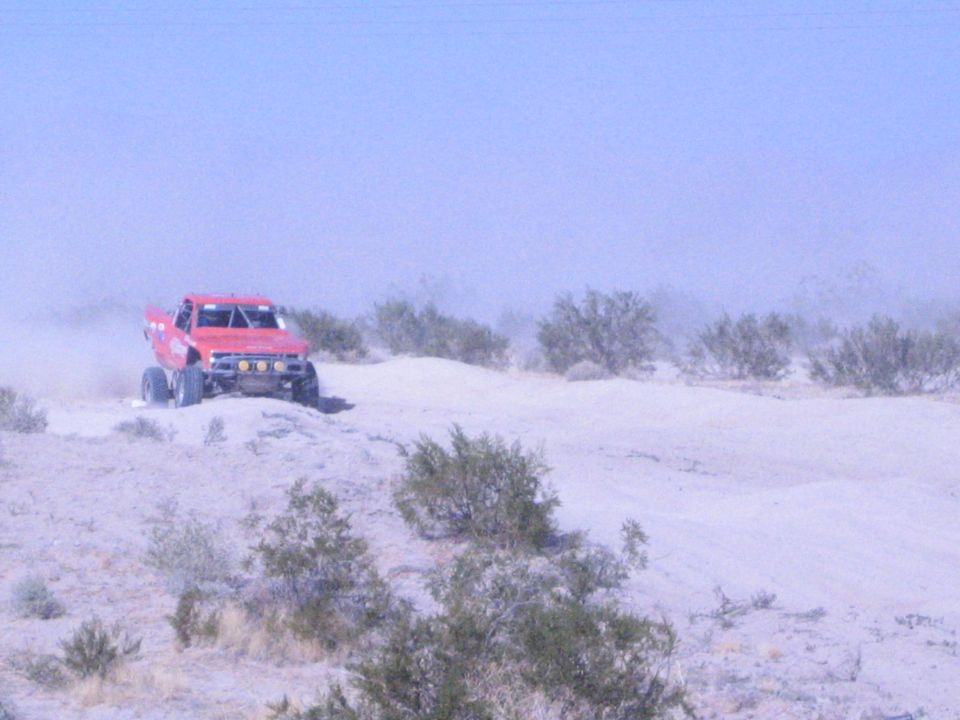 Red off-road race truck on a sandy desert berm with brush in foreground