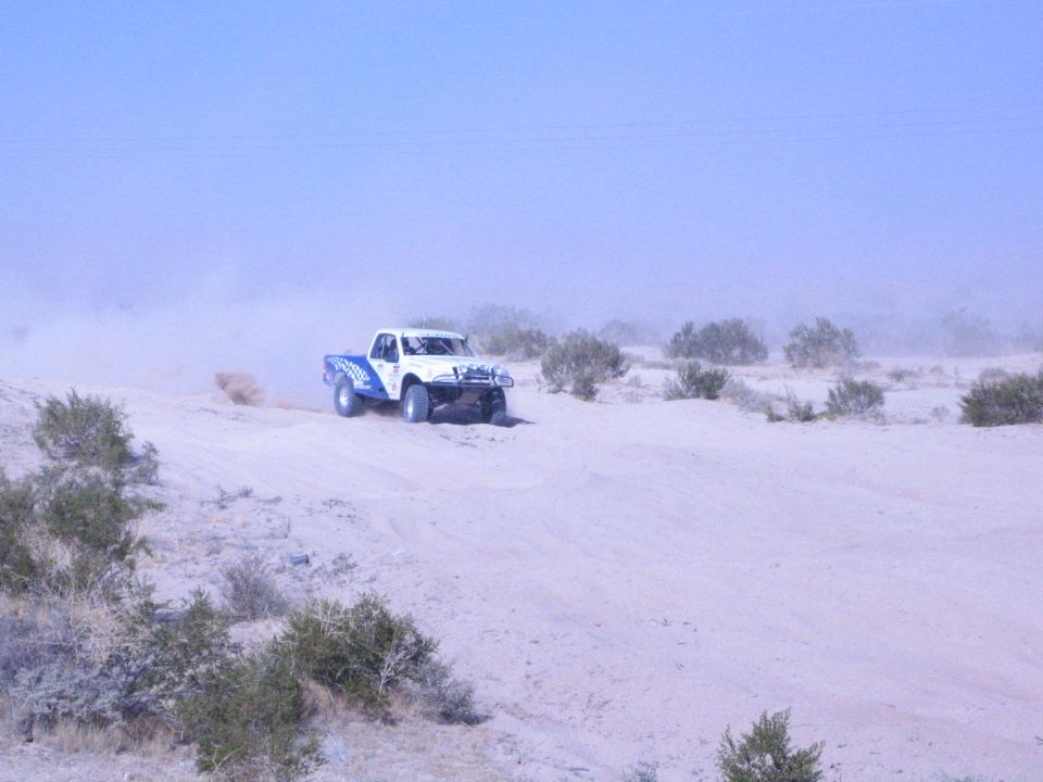 White and blue off-road race truck on pale desert sand with dust cloud