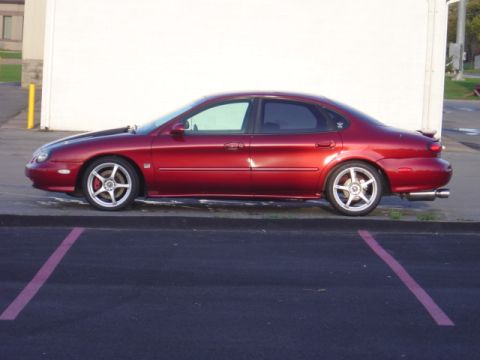 1998 Ford Taurus - A red 1998 Ford Taurus sedan is parked on asphalt, viewed from the side.