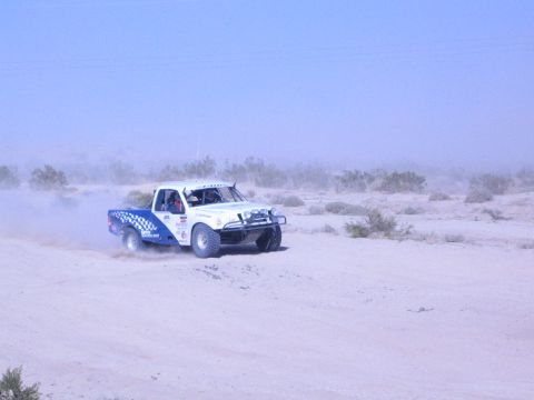 White and blue checkered race truck driving across flat desert sand