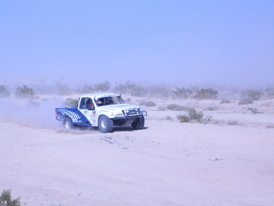 White and blue checkered race truck driving across flat desert sand