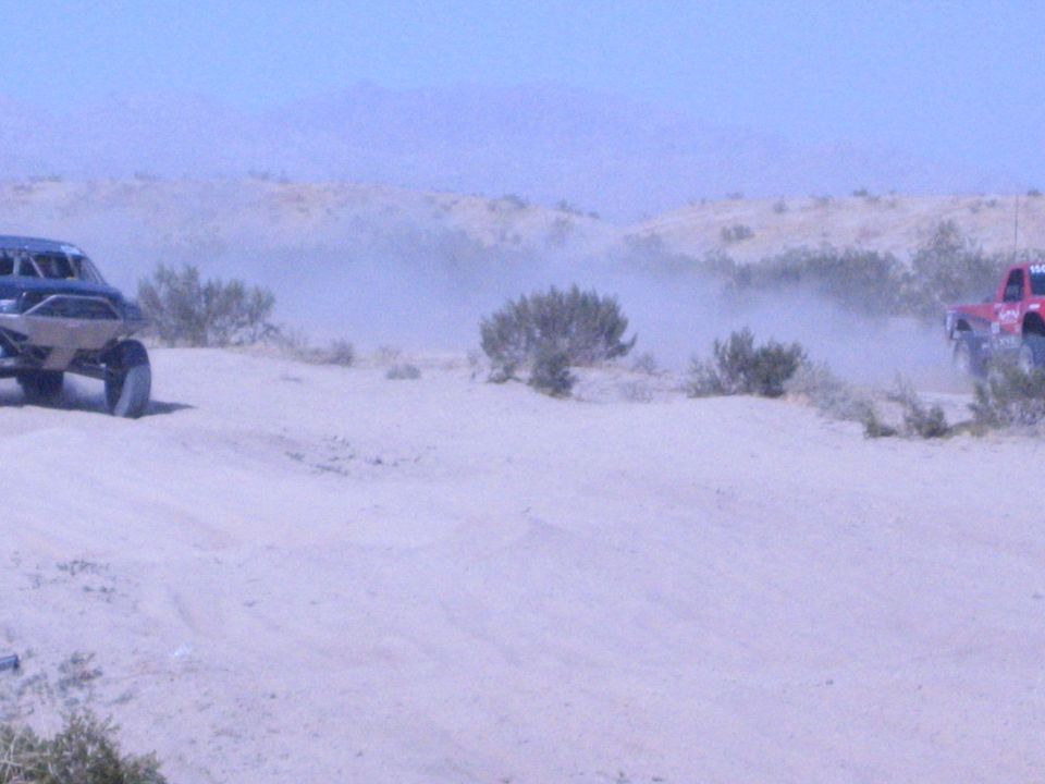 Two off-road race trucks on desert sand, one dark and one red, in dust