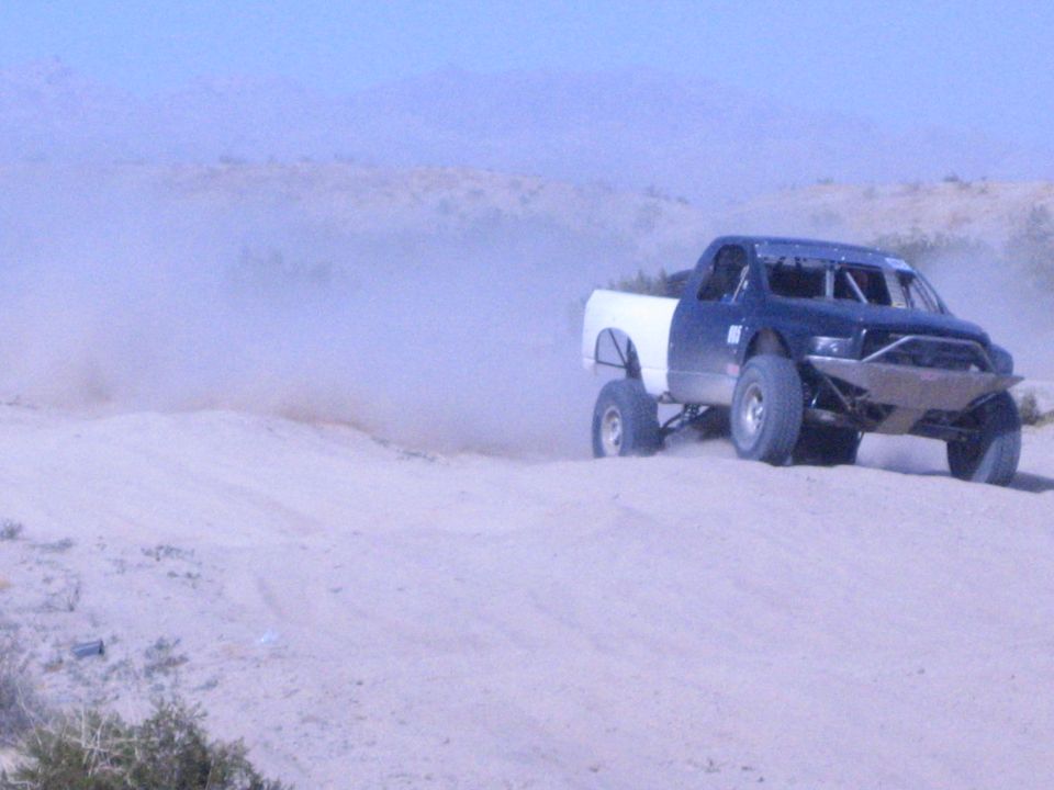 Blue race truck with white bed driving over a desert dune trailing dust