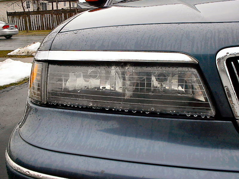 Dark blue car's front headlight assembly and chrome grille, wet with rain. A silver car is visible in the background