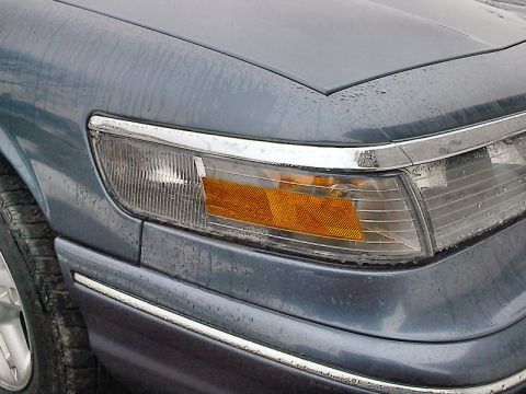 1985 Mercury Grand Marquis - Close-up of the front headlight assembly on a blue car, chrome accents and an amber turn signal indicator.