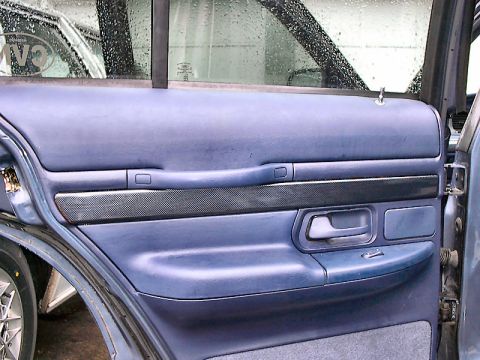 1985 Mercury Grand Marquis - The interior door panel of a car shows blue upholstery and a textured trim piece above the armrest.