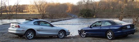 1994 Ford Probe - Two sporty coupes are parked on a gravel bank next to a river, with a bridge and trees in the background.