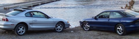 1994 Ford Probe - Two coupes, a silver Ford Mustang and a blue Ford Probe, sit parked near a waterfront.