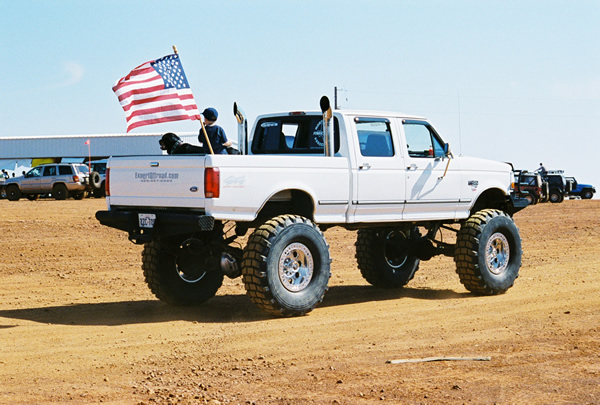 White pickup truck with oversized tires and dual exhaust pipes, flying an American flag