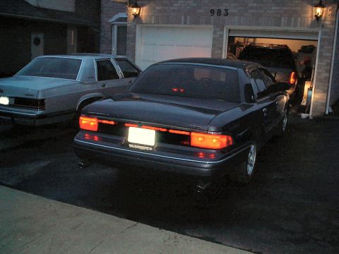 1985 Mercury Grand Marquis - Rear view of a dark sedan parked in a driveway next to another car, with a garage in the background.