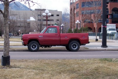 1972 Ford Ranchero - Side view of a red pickup truck parked on a city street with buildings and mountains visible.