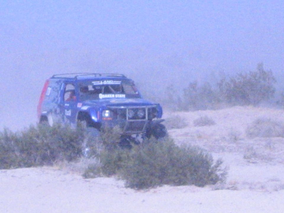 Blue off-road race SUV with red rear panel among desert brush in dust