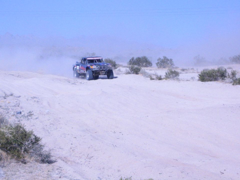 Dark off-road race truck on a desert sand rise with dust and brush