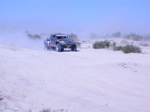 Dark blue off-road race truck driving over pale desert sand in haze