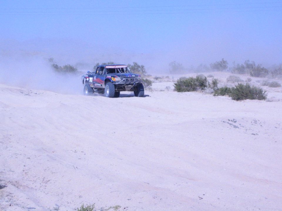 Dark blue off-road race truck driving over pale desert sand in haze