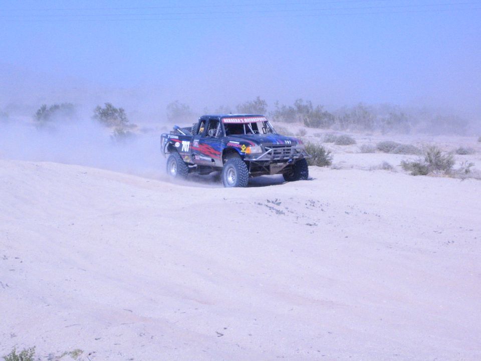 Dark blue race truck with number on desert sand throwing dust