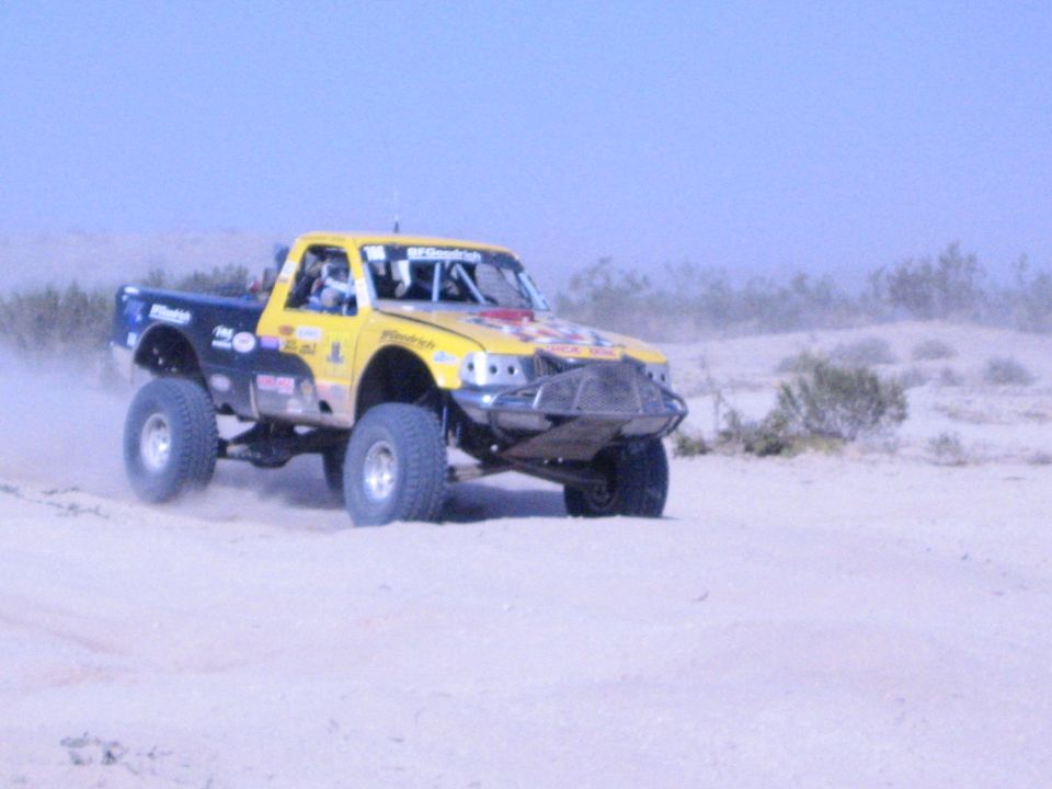 Yellow and blue tube-chassis race truck cresting a sandy desert rise