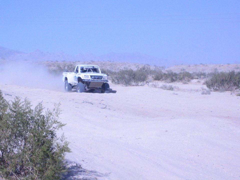 White off-road race truck driving down a wide sandy desert trail