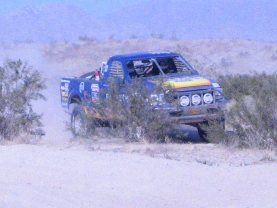 Blue off-road race truck with sunburst hood passing desert brush