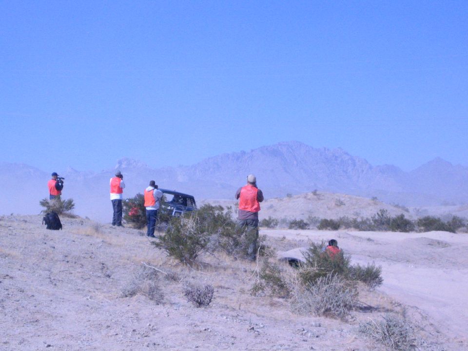Spectators in orange vests watch off-road racing in a desert valley with mountains