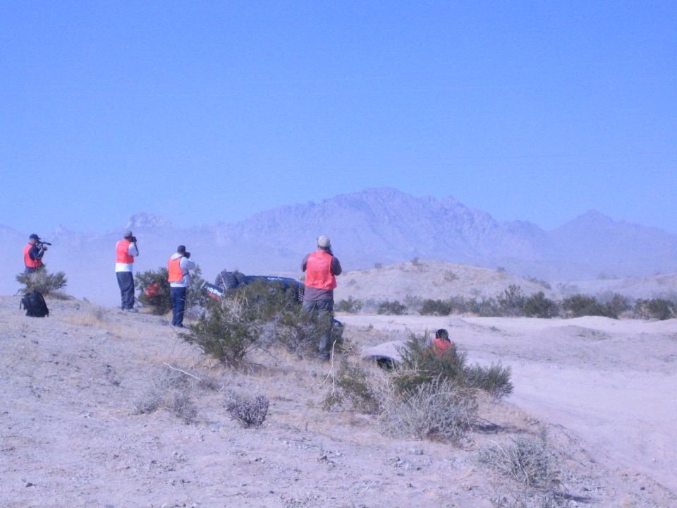 People in orange vests on a desert ridge watching distant off-road race vehicles