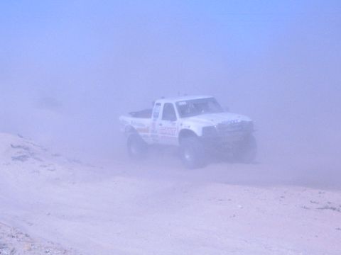 White off-road race truck partly obscured by heavy dust on a sand course