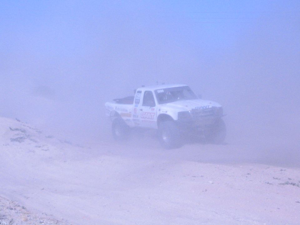 White off-road race truck partly obscured by heavy dust on a sand course
