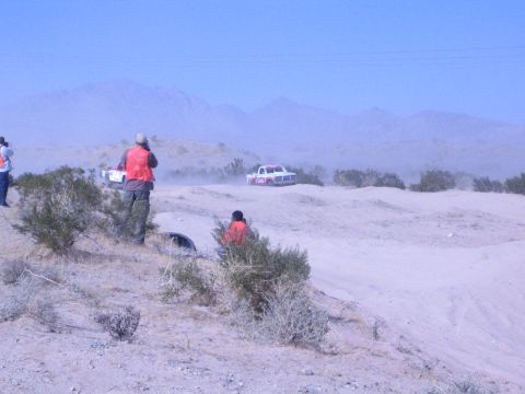 Spectators in orange vests watch race trucks across a dusty desert course