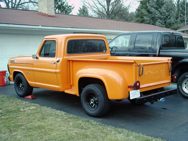 Orange Ford stepside pickup parked in driveway beside black SUV, rear three-quarter