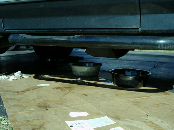 Two drain pans and a wrench on cardboard under a dark truck during a fluid change