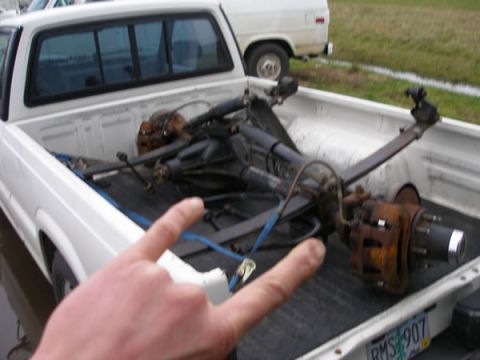 1994 Ford Bronco - Axles are loaded in the bed of a white pickup truck, with another vehicle visible in the background.