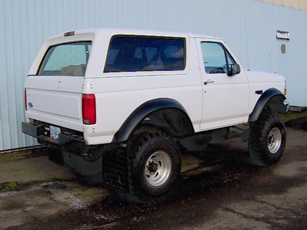 Lifted white Ford Bronco with black fender flares and large mud tires, rear three-quarter view by a blue building