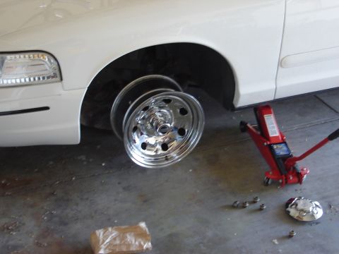 2000 Ford Crown Victoria - A white 2000 Ford Crown Victoria is shown with its front wheel removed and a red hydraulic jack in place.