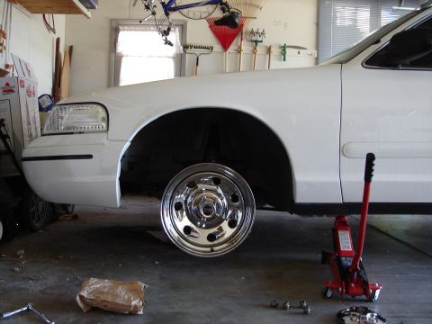 2000 Ford Crown Victoria - A white 2000 Ford Crown Victoria is shown in a garage with its front wheel removed and the vehicle supported by a jack.