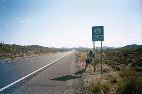 1990 Buick Park Avenue - A person pauses by a highway sign marking the entrance to Peoria, Arizona, with desert scenery in the background.