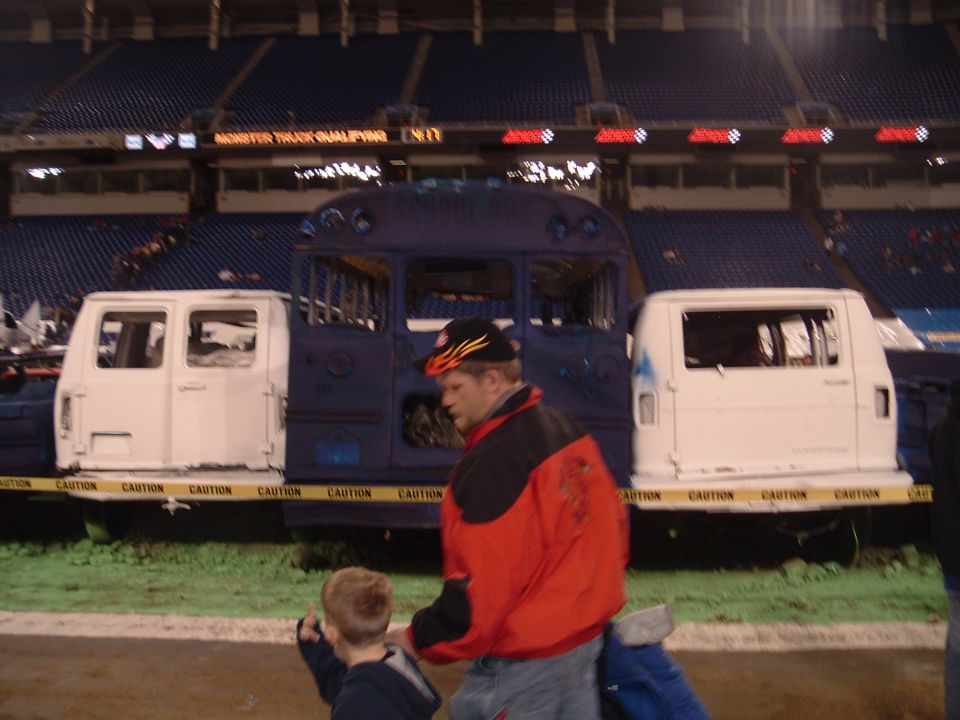 Crushed blue school bus and white vans behind caution tape at an indoor monster truck event, man and boy walking past