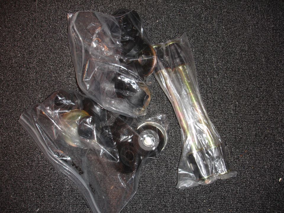 Several black and silver automotive suspension components in plastic bags on a dark carpeted surface